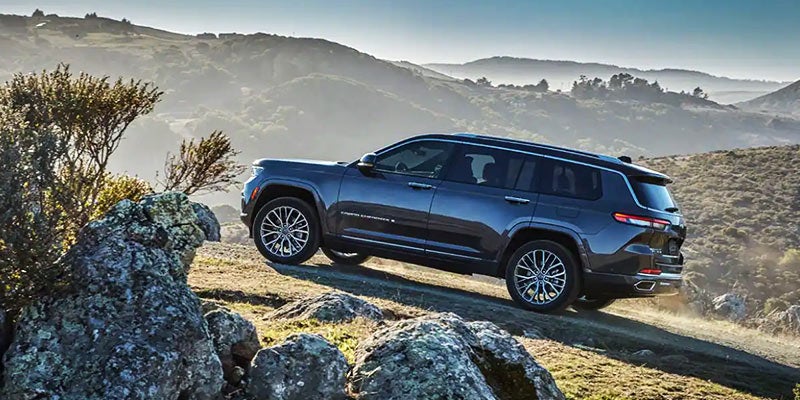 Dark blue Jeep Grand Cherokee L ascending a dirt trail, with mountains and trees in the misty background.