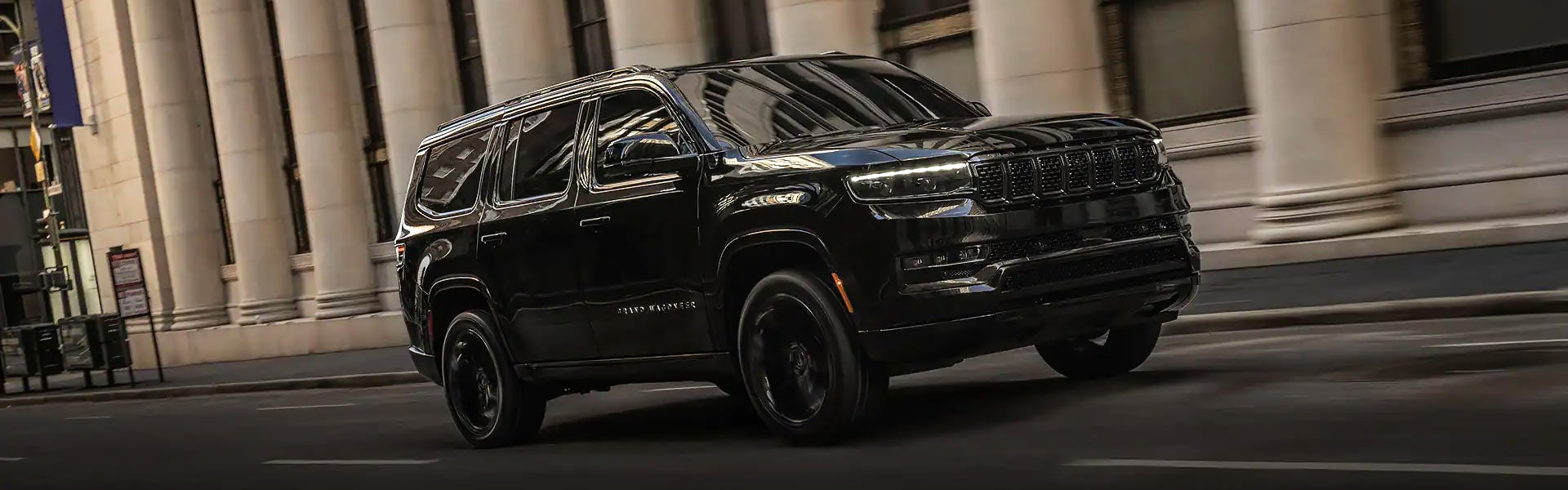 A shiny black SUV is driving on a city street past a building with large ornate columns.