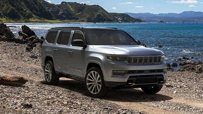 A silver Jeep Wagoneer is parked on a rocky shoreline next to a bright blue ocean with mountains in the background.