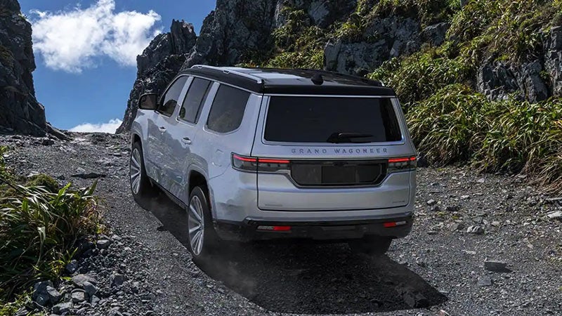 Rear view of a silver Jeep Grand Wagoneer driving up a rocky mountain trail among tall rocks and green plants.