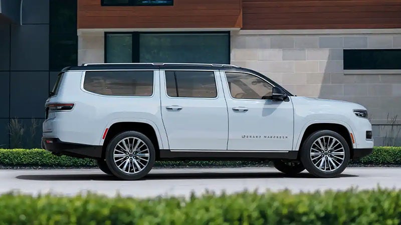 Side profile of a white 2022 Grand Wagoneer SUV parked on a paved driveway in front of a modern building.