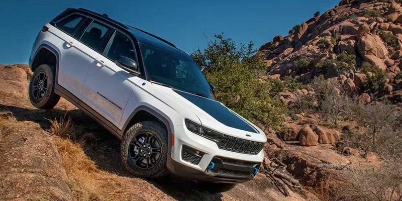 A white 2025 Jeep Grand Cherokee climbs a rocky off-road trail against a clear blue sky.