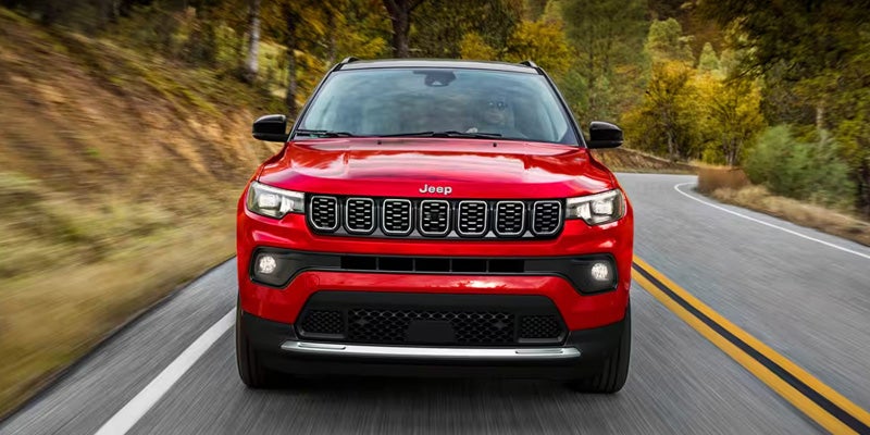 Front view of a red Jeep Compass driving on a winding, tree-lined road in autumn.