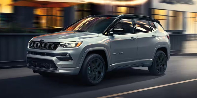 A silver Jeep Compass with a black roof drives on a city street at dusk.