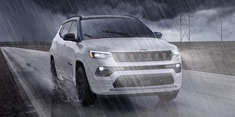 A white Jeep Compass driving on a wet road in heavy rain under a dark, cloudy sky.