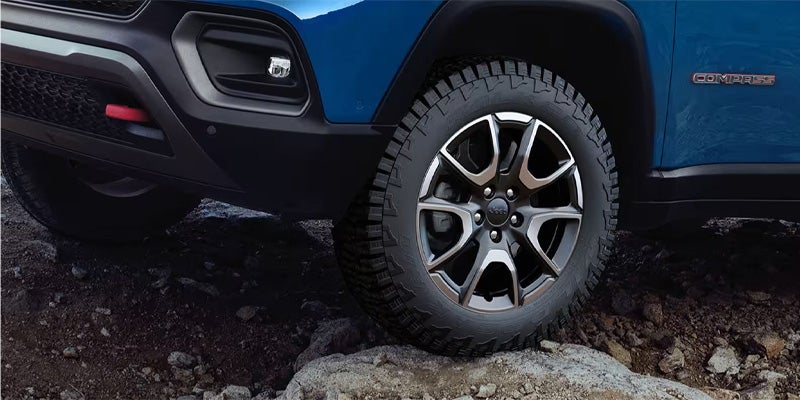 Front quarter view of a blue Jeep Compass on a rocky, dark ground. Its tire has large treads, and the rim is gray and silver.