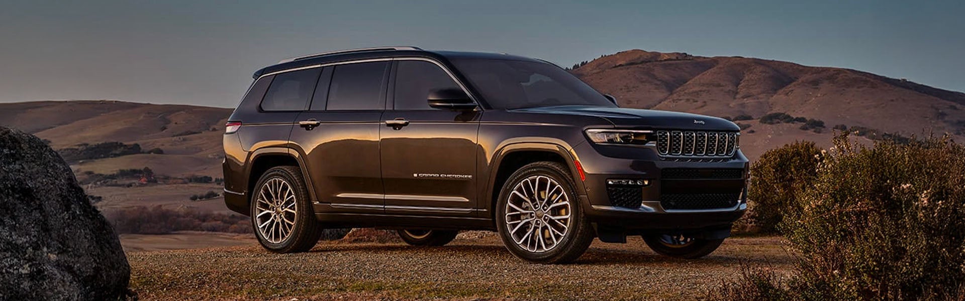 A dark grey Jeep Grand Cherokee SUV is parked on a rocky hill with mountains in the background.
