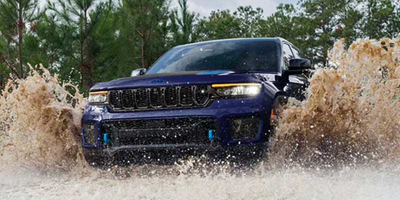 A dark blue Jeep Grand Cherokee 4xe drives through muddy water, creating huge splashes. Trees are visible in the background.