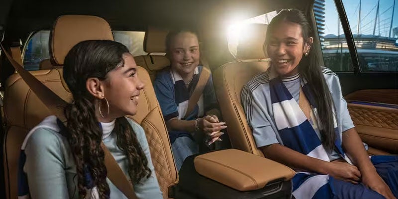 Three smiling young women ride in a car, two wearing blue and white striped shirts and one in a light blue sweatshirt.