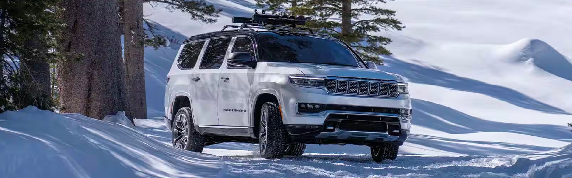 A white Jeep Grand Wagoneer with skis on the roof rack drives through a snowy, mountainous landscape.