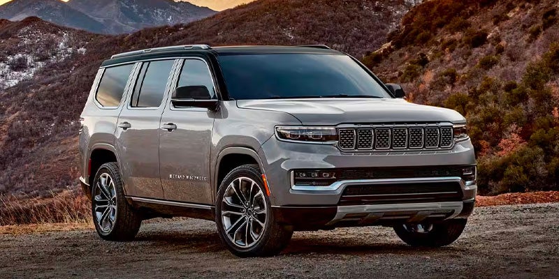 A silver Jeep Grand Wagoneer L SUV parked on a dirt road with mountains and sky in the background at sunset.
