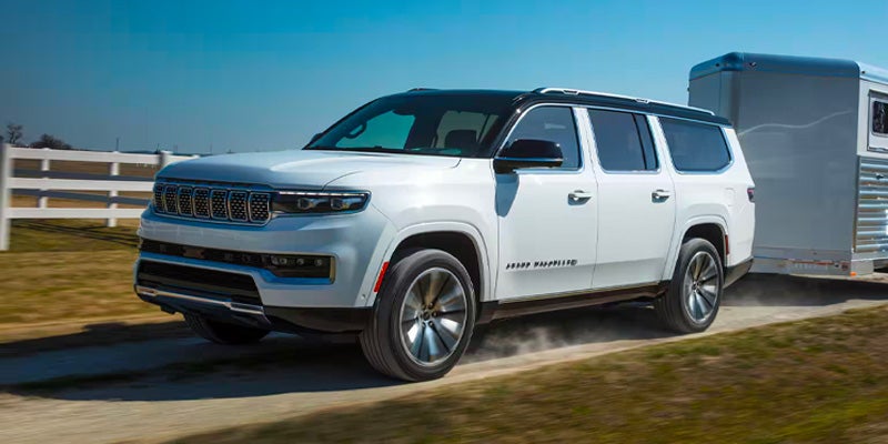 Front 3/4 view of a white 2022 Grand Wagoneer towing a silver horse trailer on a dirt road.