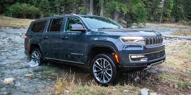 A dark blue Wagoneer SUV drives through a shallow river, with water splashing around its wheels.