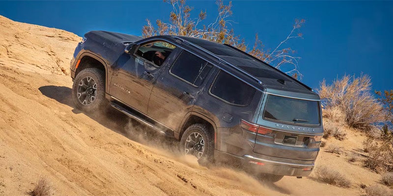 A gray Jeep Wagoneer SUV drives down a dusty, sandy hill in an arid landscape under a clear blue sky.