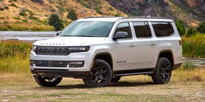 A white 2024 Jeep Grand Wagoneer SUV parked on dirt next to a body of water and treeline.