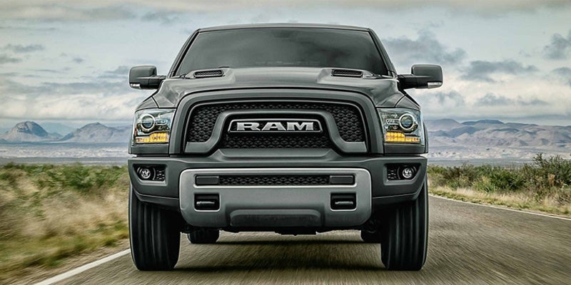 A dark gray Ram truck with an aggressive grill and shiny headlights drives on a rural road under a gray sky.