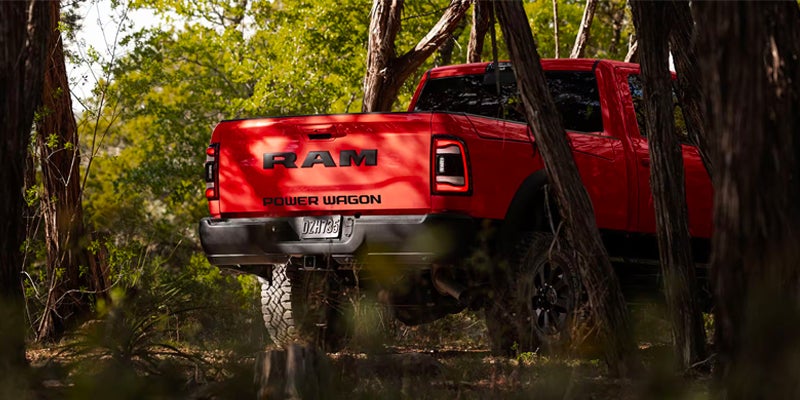 Rear view of a red Ram Power Wagon pickup truck parked in a dense forest, with tree trunks in the foreground.
