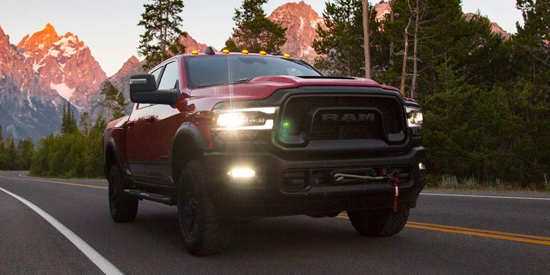 A red Ram Rebel truck drives on a mountain road with its headlights on, mountains and trees in the background.
