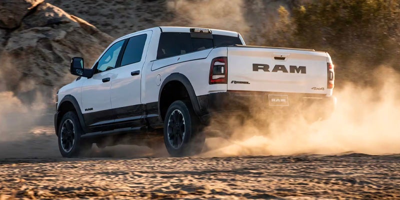 A bright white Ram Rebel truck kicks up dust on a sandy, off-road track, seen from the rear passenger side.