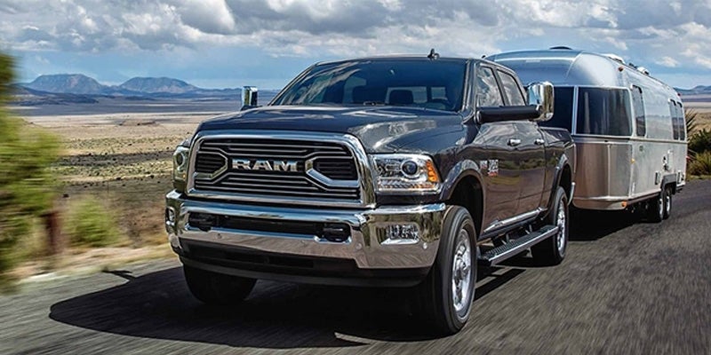 A grey Ram truck towing a silver Airstream trailer on a highway through a desert landscape. Mountains are in the background under a cloudy sky.