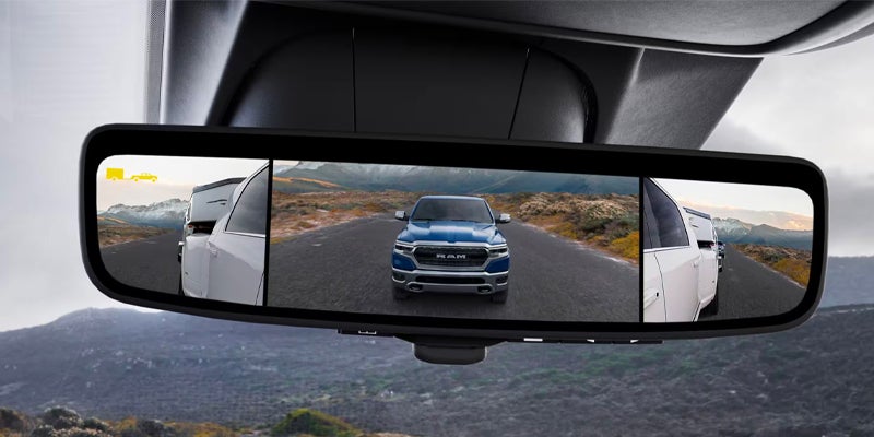 Digital rearview mirror shows a blue truck on a road, flanked by parts of a white truck and trailer.