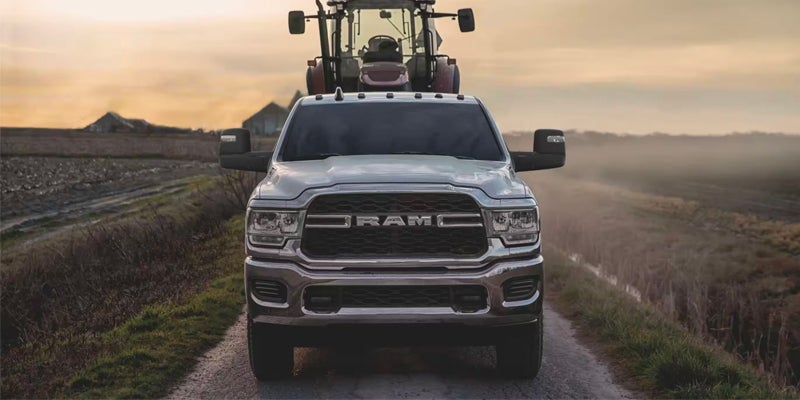 A white Ram truck faces forward on a dirt road, with a large tractor directly behind it. A farm field and buildings are in the background under a hazy sky.