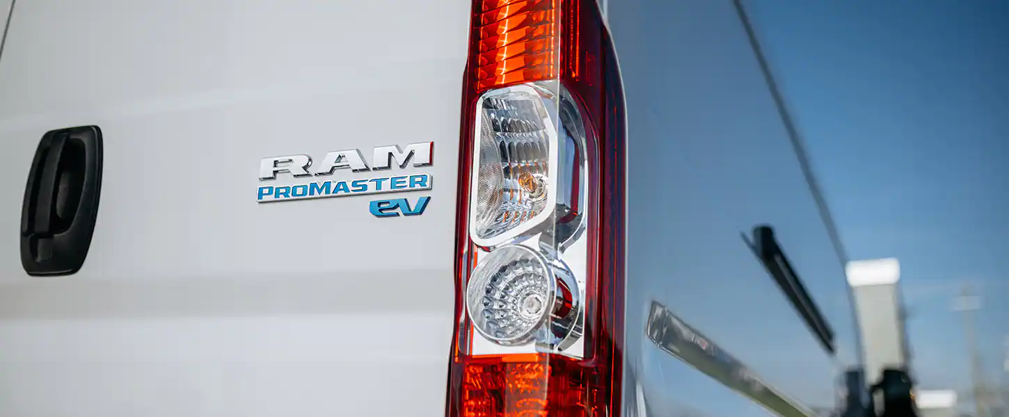 Close-up of a white van showing "RAM ProMaster EV" badge in blue and silver, next to a red and clear taillight.