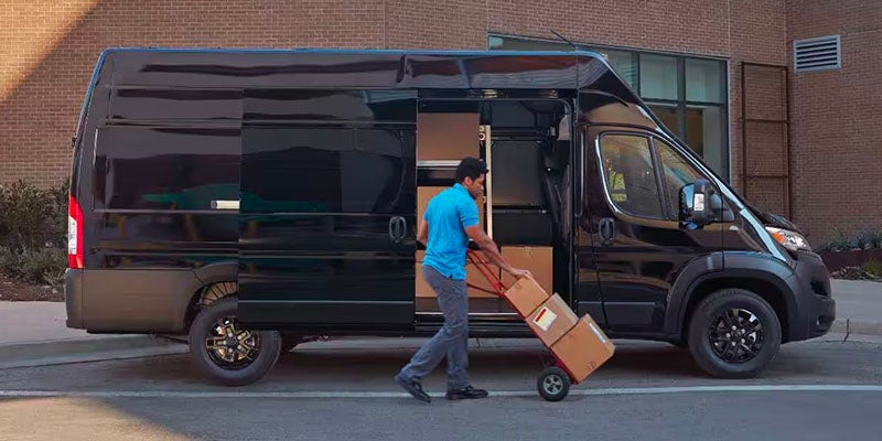 A delivery driver uses a hand truck to load brown boxes into the open side door of a black delivery van.
