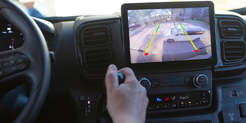 A hand reaches for the gear selector inside a vehicle. A large screen displays a rearview camera view with a white car in the driveway.